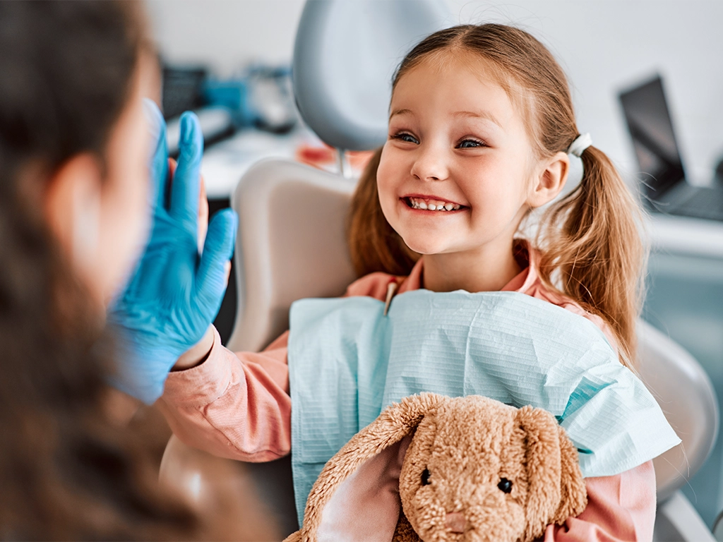 child sitting in a dental chair holding a toy rabbit and cheerfully giving a high-five to the dentist